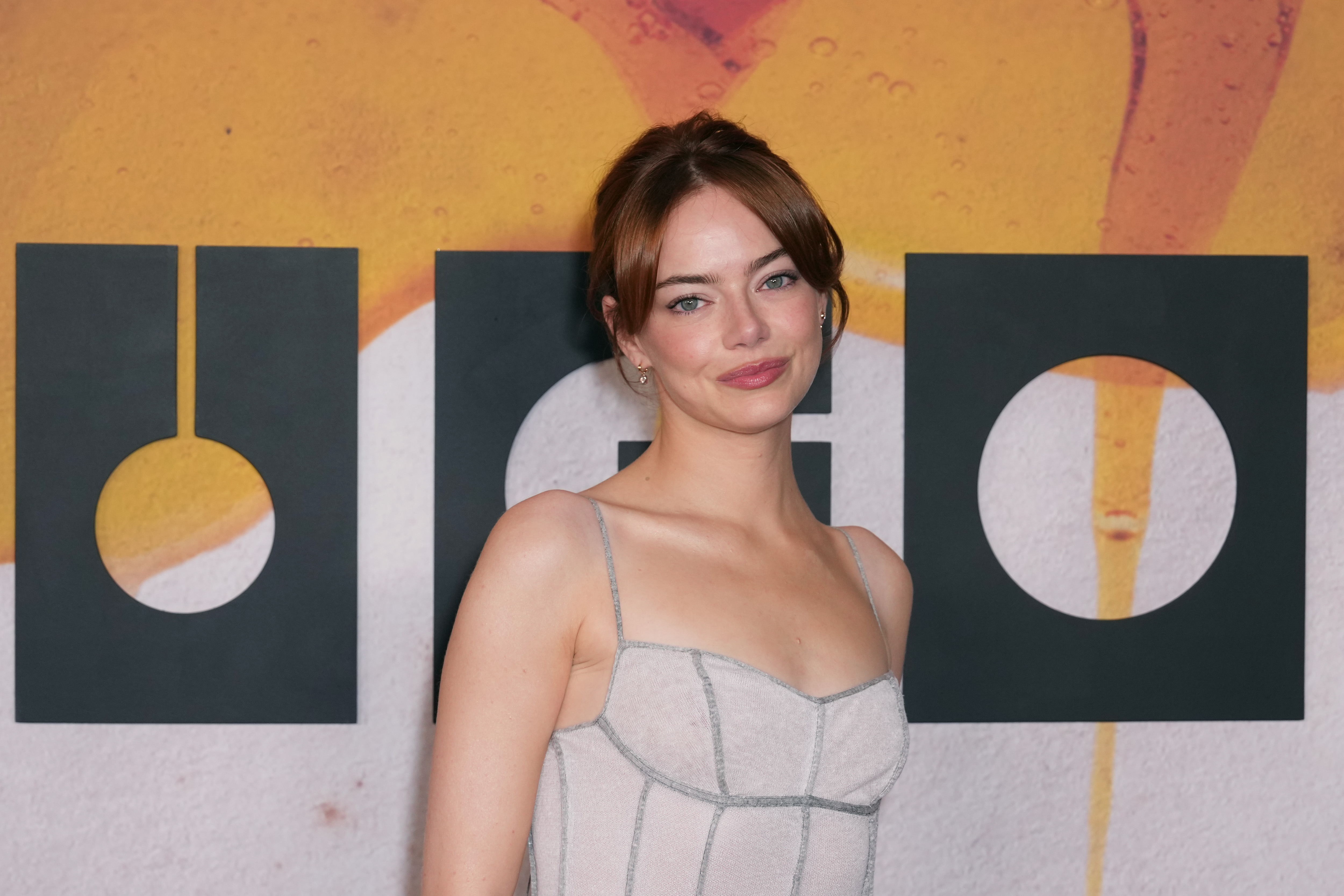 Emma Stone pose avec assurance, les épaules inclinées et les lèvres esquissant un sourire complice. La douce brillance de sa robe capte la lumière sur le fond audacieux « Bugonia ».