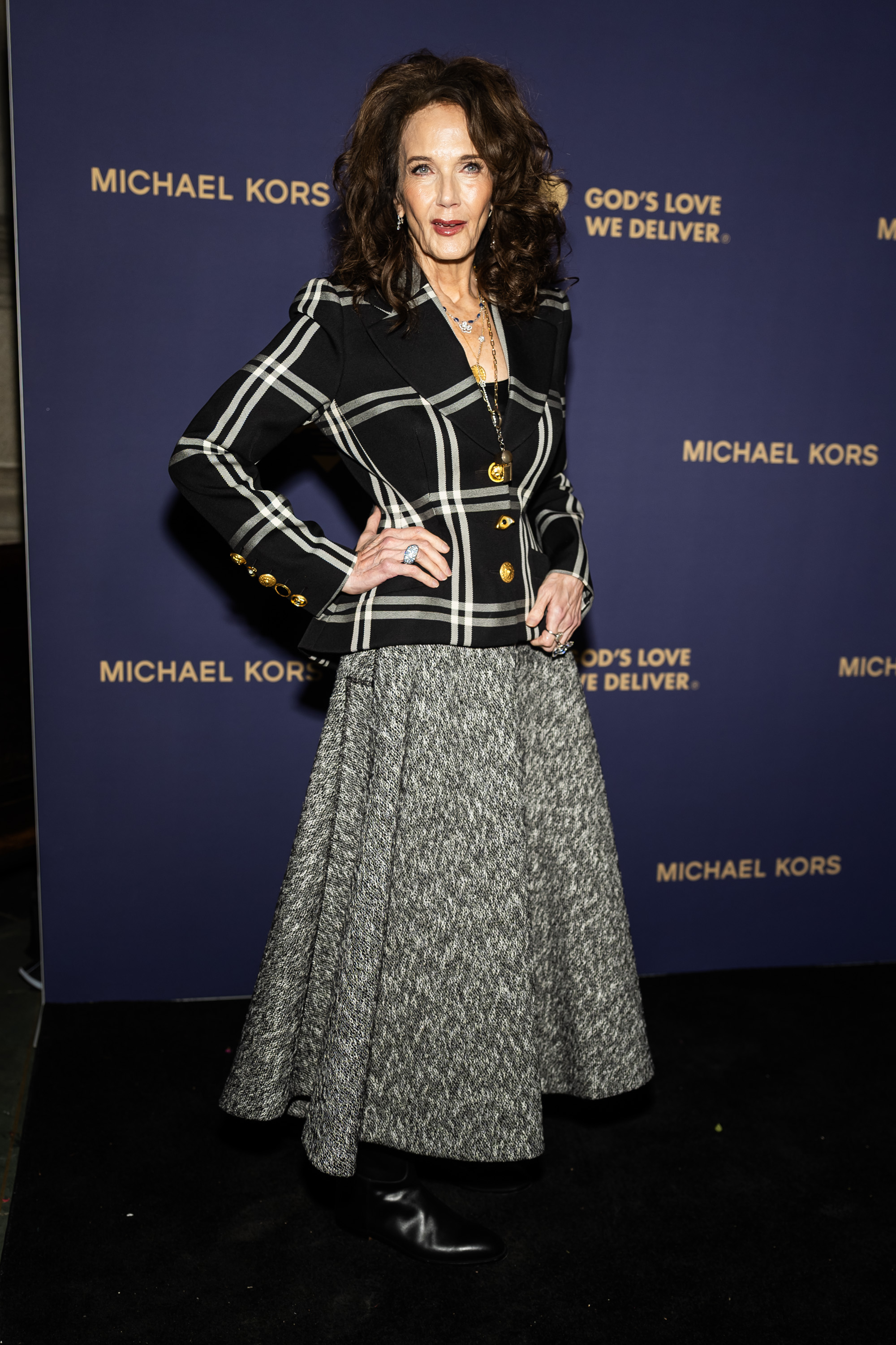 Lynda Carter pose sur le tapis rouge lors de l'événement God's Love We Deliver dans une superbe tenue noir et blanc. Elle associe un blazer à carreaux ajusté avec des boutons dorés audacieux et une jupe grise texturée, complétant son look avec des bijoux imposants, des bottes en cuir et des boucles volumineuses.