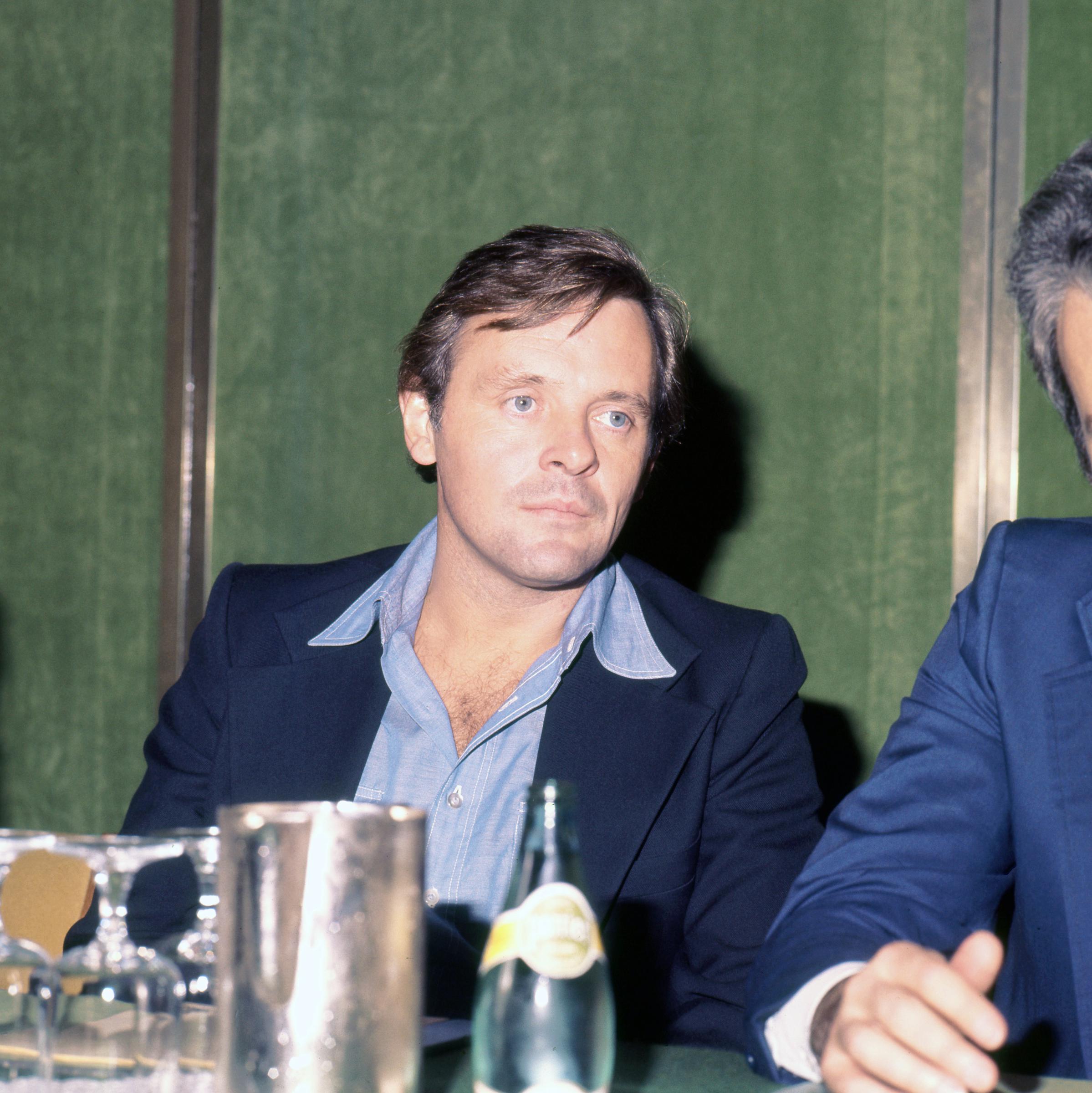 Assis avec assurance à une table de conférence de presse à Londres, Anthony Hopkins affiche un calme qui laisse deviner une introspection tranquille. Vêtu d'une chemise bleue impeccable et d'une veste sur mesure, il rayonne d'une sophistication naturelle. L'éclairage tamisé et les vitres réfléchissantes confèrent à la scène une atmosphère intemporelle de sérénité et d'élégance.