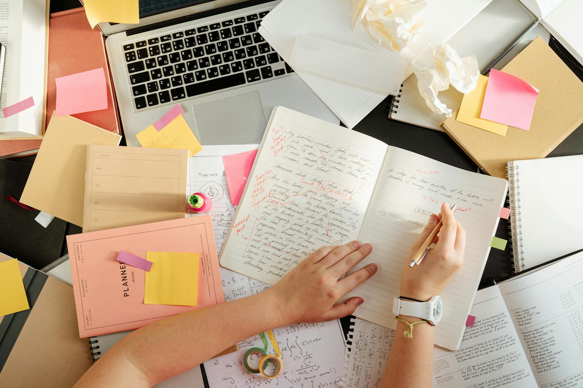 Un bureau en désordre avec des livres, un ordinateur portable et une personne qui écrit dans un carnet | Source : Pexels