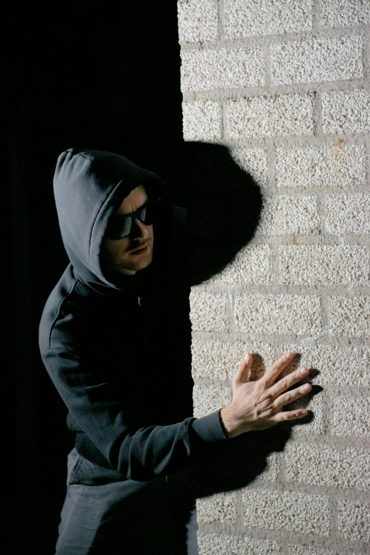 Un homme suspect portant un sweat à capuche et des lunettes de soleil se tient derrière un mur | Source : Pexels