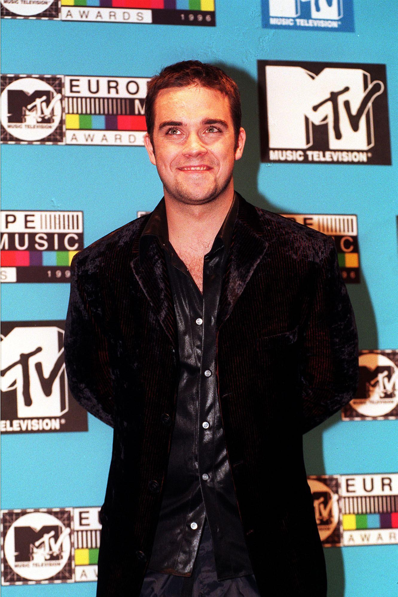 Robbie Williams assiste aux MTV Europe Music Awards en 1996, vêtu d'un blazer en velours et d'une chemise en satin. Cette apparition marque l'un des premiers tournants de sa carrière solo, alors qu'il commence à sortir de l'ombre de Take That et à tracer sa propre voie dans la musique pop.