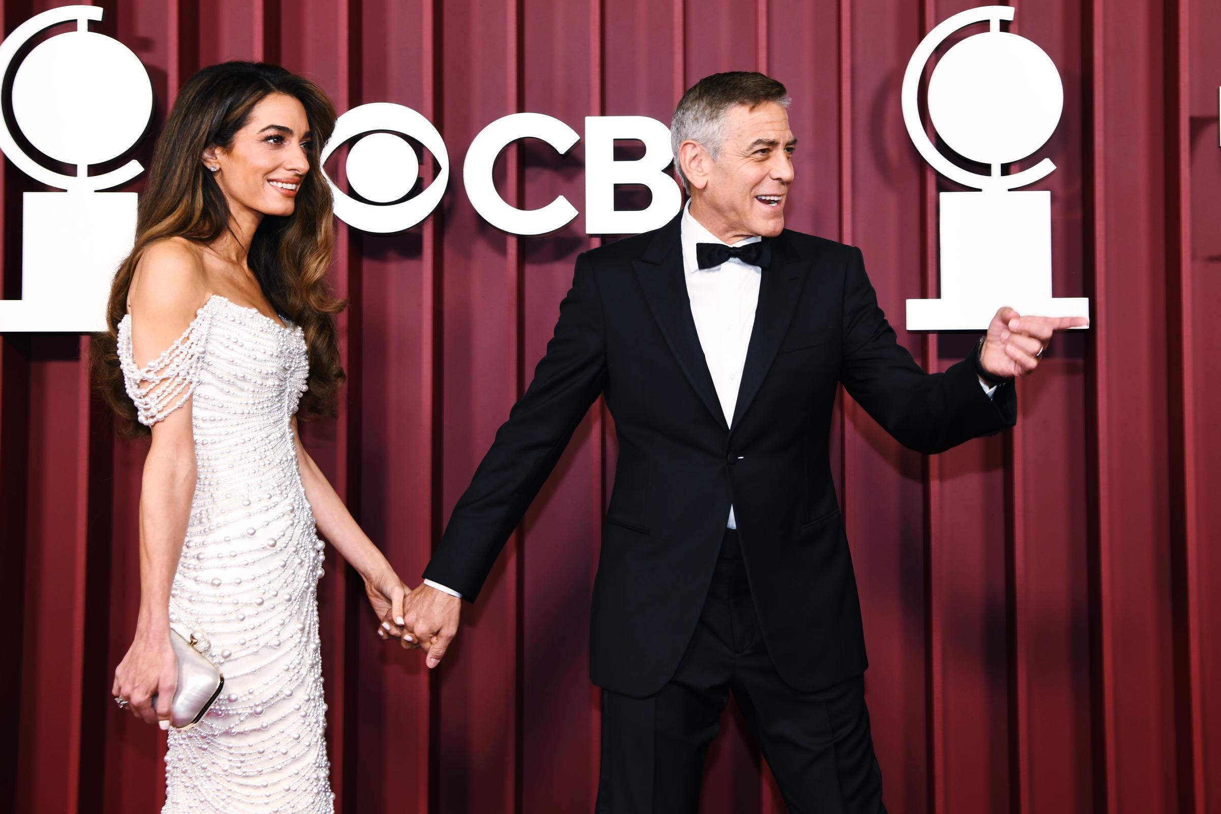 Amal Clooney rayonne tandis que George Clooney discute de façon expressive, leurs doigts entrelacés sur le tapis rouge. Sa robe ornée de perles éblouit sous les lumières, tandis qu'il entretient l'ambiance avec un geste enjoué.