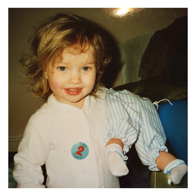 Rosie Alice Huntington-Whiteley sourit sur un cliché d'enfance, portant une tenue blanche avec un badge "J'ai 2 ans aujourd'hui", et tenant une poupée. | Source : Instagram/rosiehw