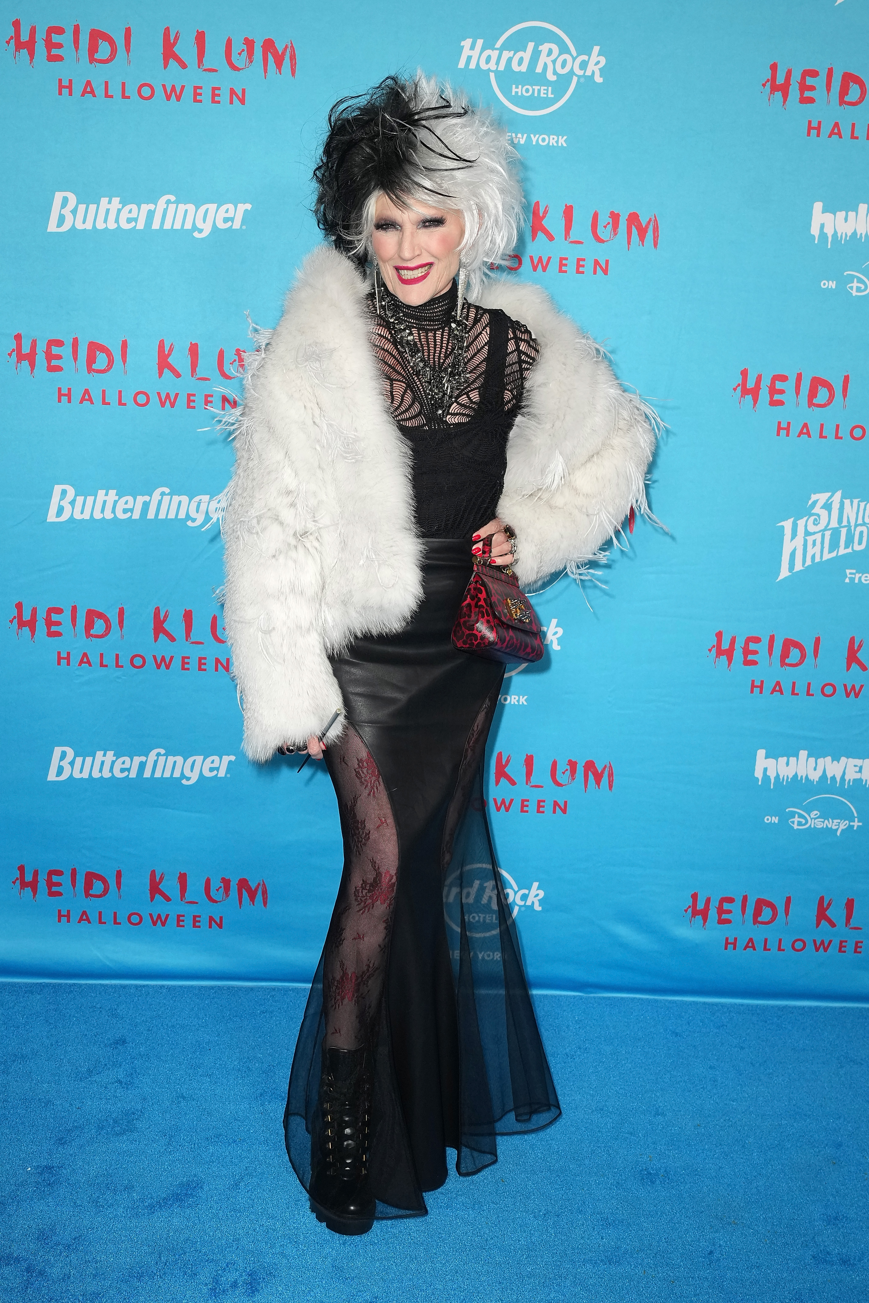 Lors de la 24e fête annuelle d'Halloween organisée par Heidi Klum, Maye Musk, la mère d'Elon Musk, a adopté un look glamour inspiré de Cruella d'Enfer. Elle portait une volumineuse perruque noire et blanche, un manteau de fourrure spectaculaire orné de plumes et une robe noire transparente superposée à des collants imprimés. La tenue était complétée par des bottes à lacets à semelles compensées, un sac à main rouge à imprimé animalier et un collier imposant, pour un look haute couture avec une touche espiègle.