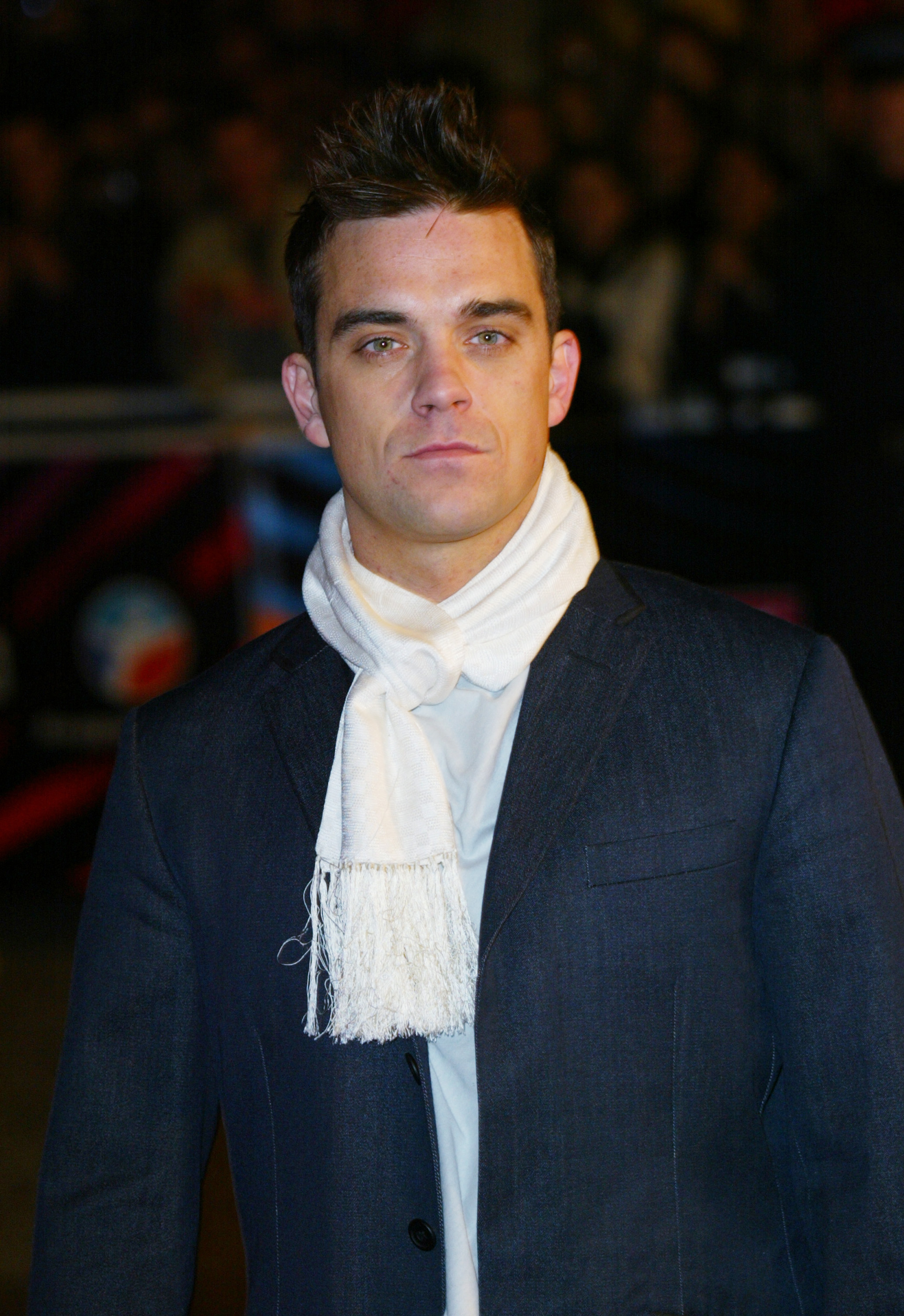 Robbie Williams aux NRJ Music Awards à Cannes, en France, en 2003, vêtu d'un costume sur mesure et d'une écharpe blanche. Avec son look élégant et son expression sérieuse, il incarne la confiance raffinée d'une pop star au sommet de sa gloire internationale.