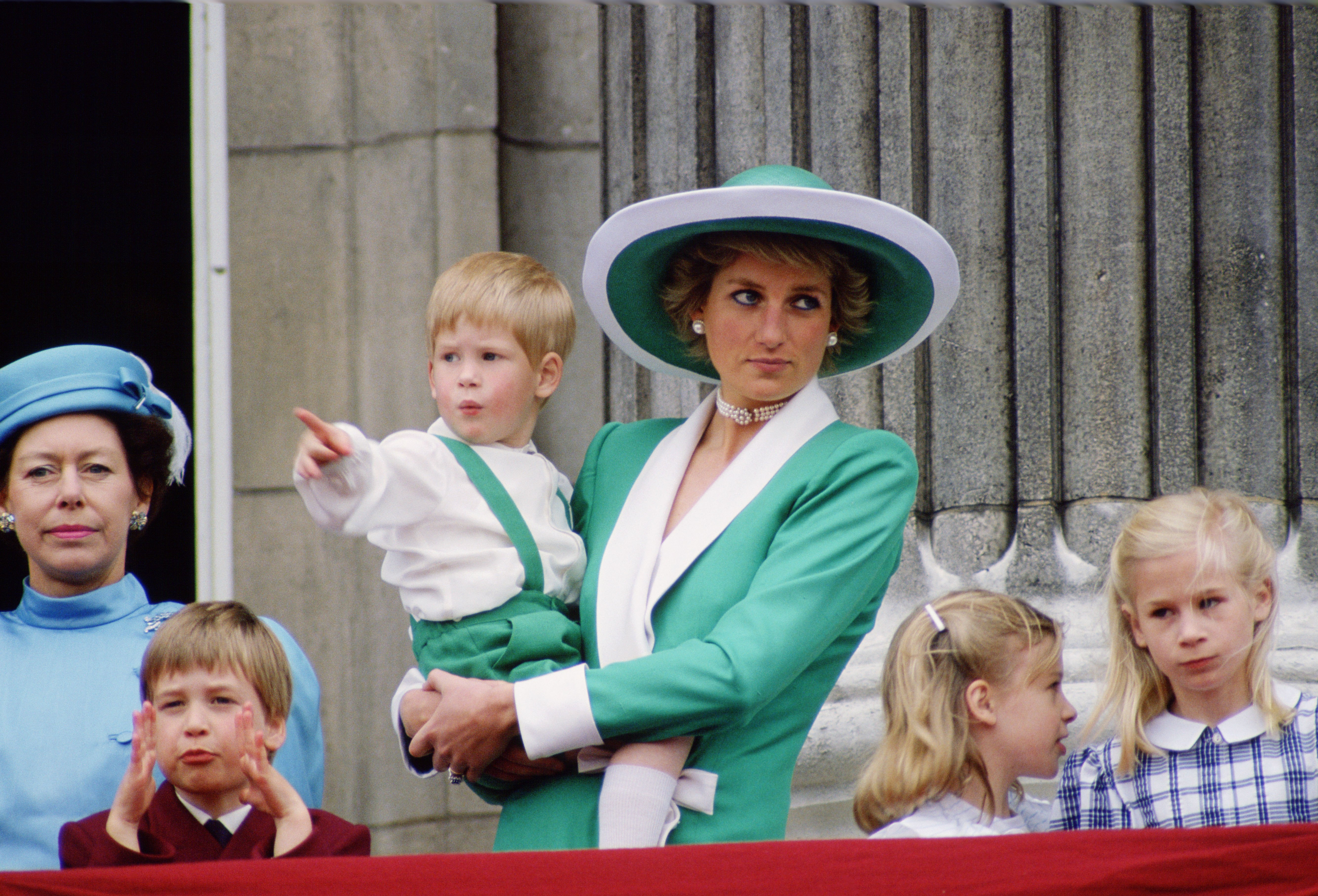 La princesse Diana, vêtue d'un tailleur vert et blanc assorti à son chapeau, tient dans ses bras le jeune prince Harry alors qu'elle assiste au défilé Trooping the Colour de 1988 depuis le balcon du palais de Buckingham. Entourée du prince William, de Lady Rose Windsor, de Lady Davina Windsor et de la princesse Margaret, la présence sereine de Diana immortalise un moment intemporel de la famille royale, encadré par la tradition et le faste.