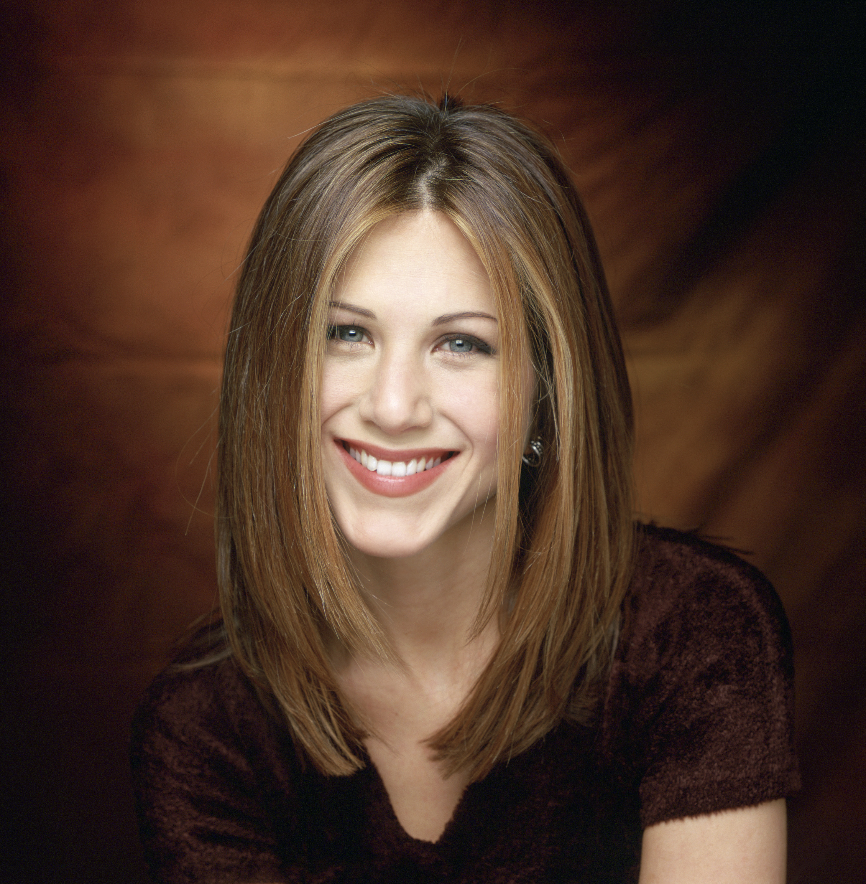Jennifer Aniston, dans le rôle de Rachel Green, sourit chaleureusement avec sa coiffure emblématique, lisse et dégradée. Elle porte un haut sombre sur un fond brun légèrement éclairé, rayonnant de charme et de confiance.