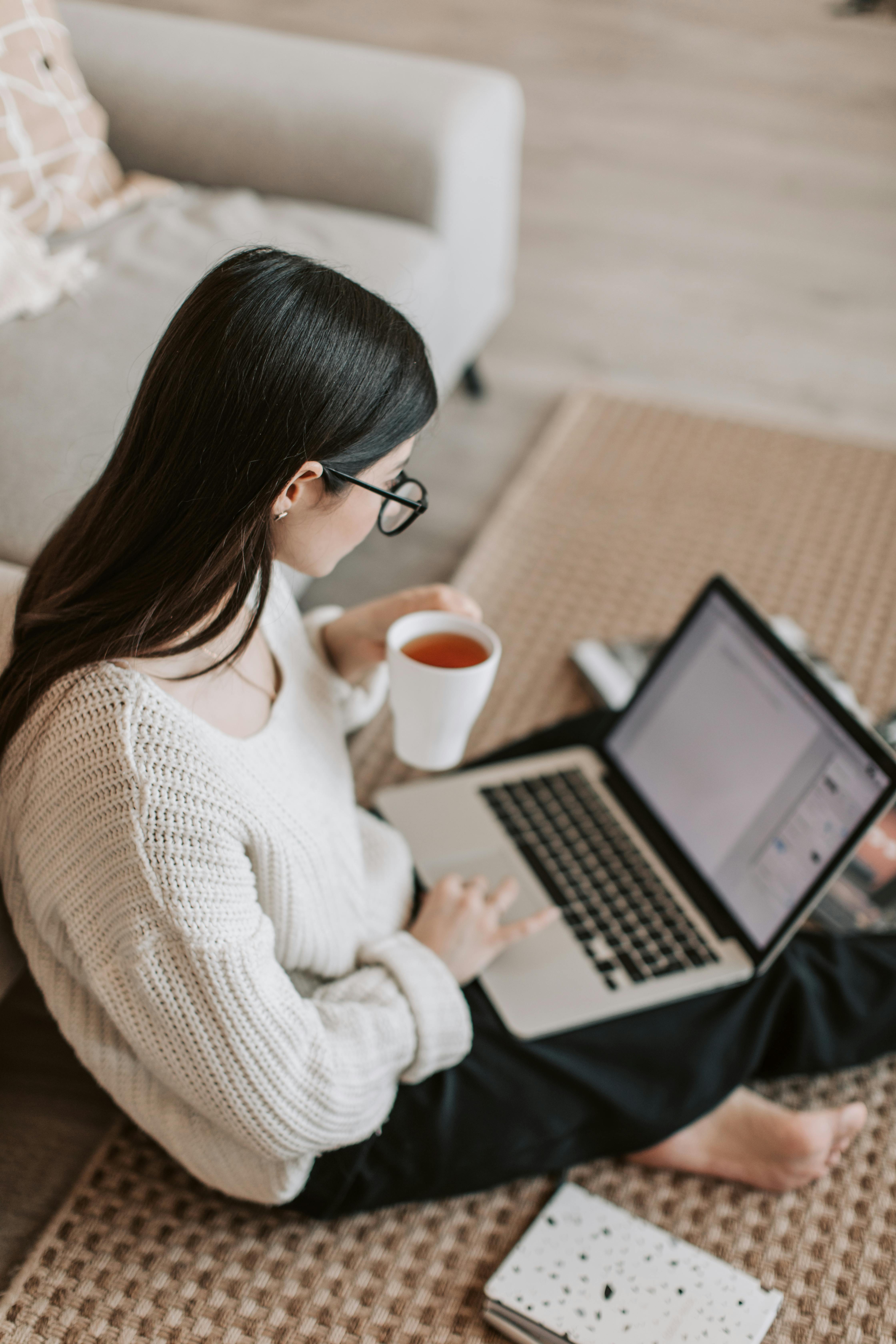 Une femme assise sur le sol et utilisant un ordinateur portable tout en tenant une tasse de boisson | Source : Pexels