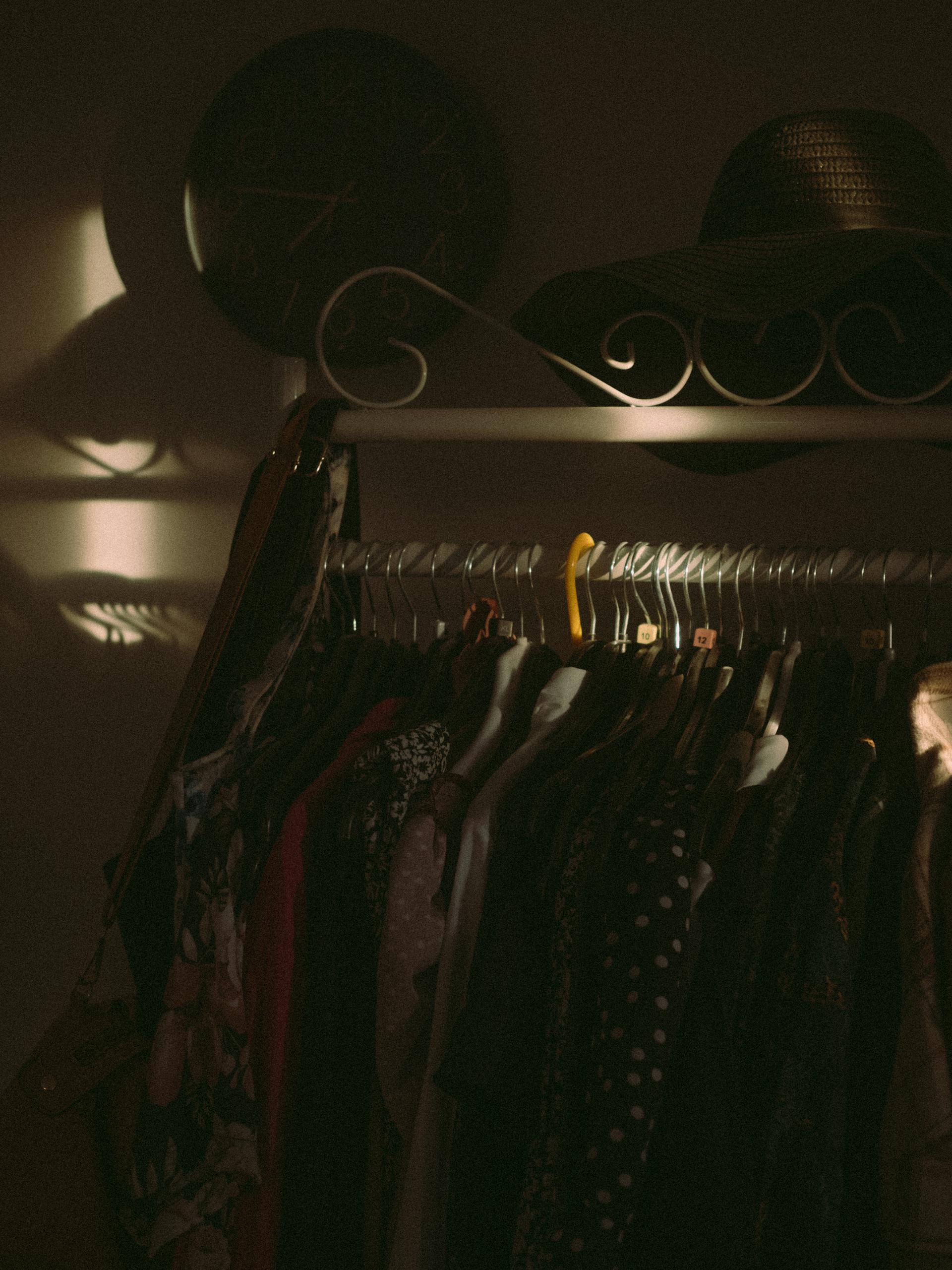 Une photo sombre de vêtements suspendus à une étagère avec un chapeau sur le dessus et une horloge accrochée au mur | Source : Pexels