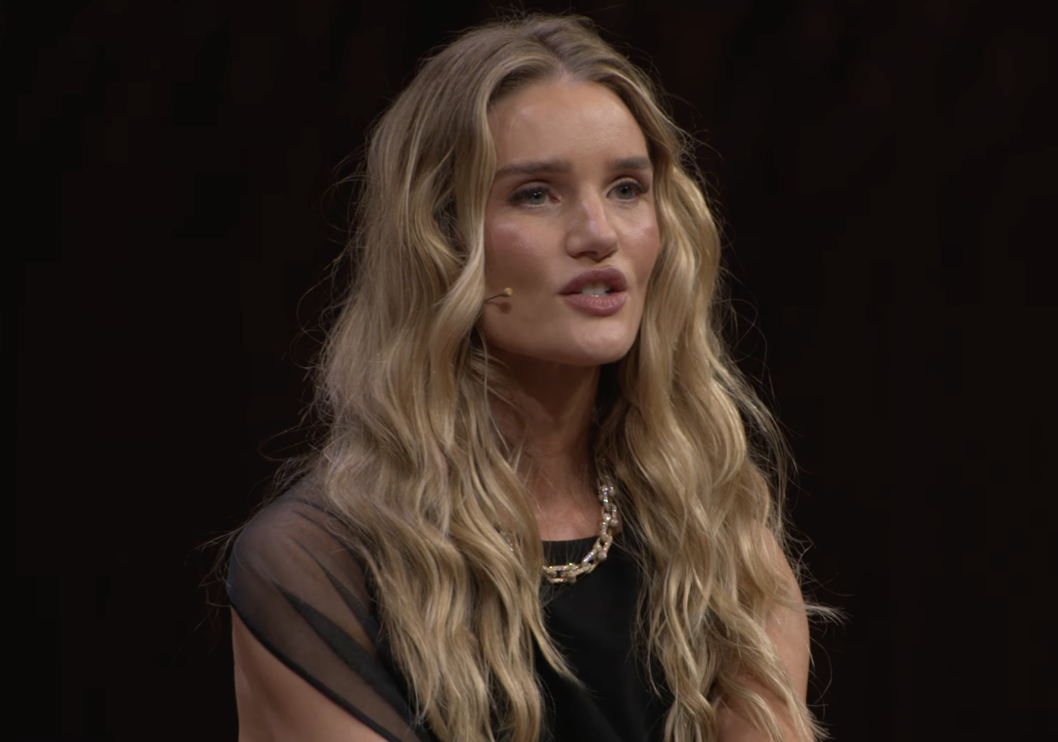 Rosie Huntington-Whiteley apparaît sur scène en plein discours, portant une robe noire élégante avec des détails transparents et un collier en or. | Source : YouTube/VogueAustralia