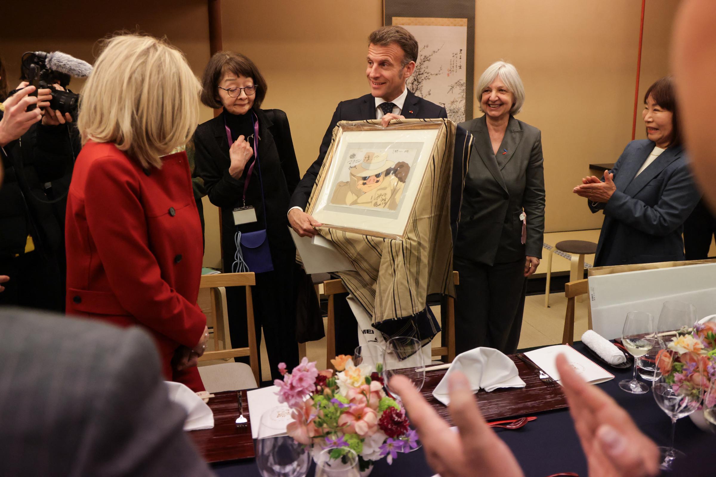 Le président français Emmanuel Macron (au centre) montre un cadeau qu'il a reçu de l'animateur, cinéaste et mangaka japonais Hayao Miyazaki (absent de la photo), aux côtés de l'ambassadrice de France au Japon, Béatrice Le Fraper du Hellen (au centre à droite), lors d'un dîner avec son épouse Brigitte Macron (à gauche) au Happo-en à Tokyo, le 31 mars 2026 I Source : Getty Images