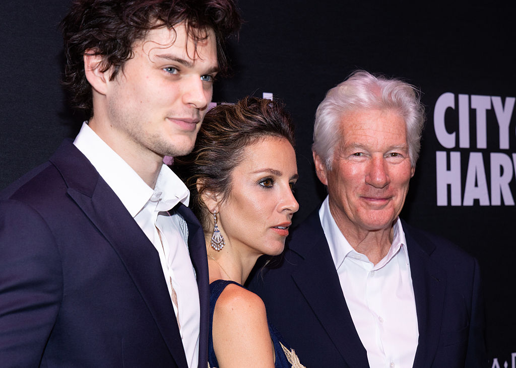 Au gala City Harvest, Homer, Alejandra et Richard Gere se tiennent côte à côte, faisant face aux caméras avec une élégance posée, chacun dégageant une confiance tranquille et une prestance digne du tapis rouge sur fond raffiné.