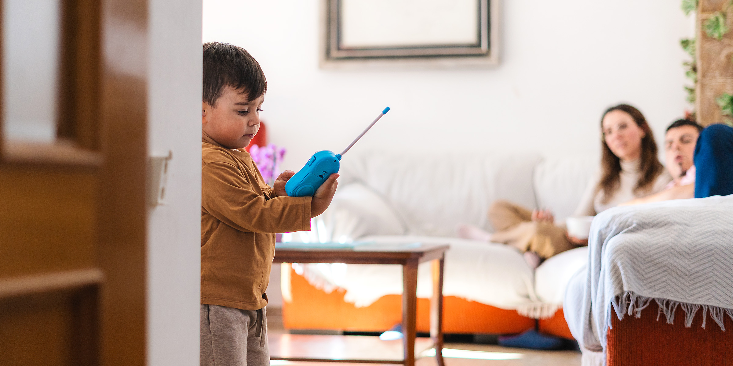 Un petit garçon qui joue avec un talkie-walkie bleu pendant que deux adultes le regardent depuis le canapé | Source : Shutterstock