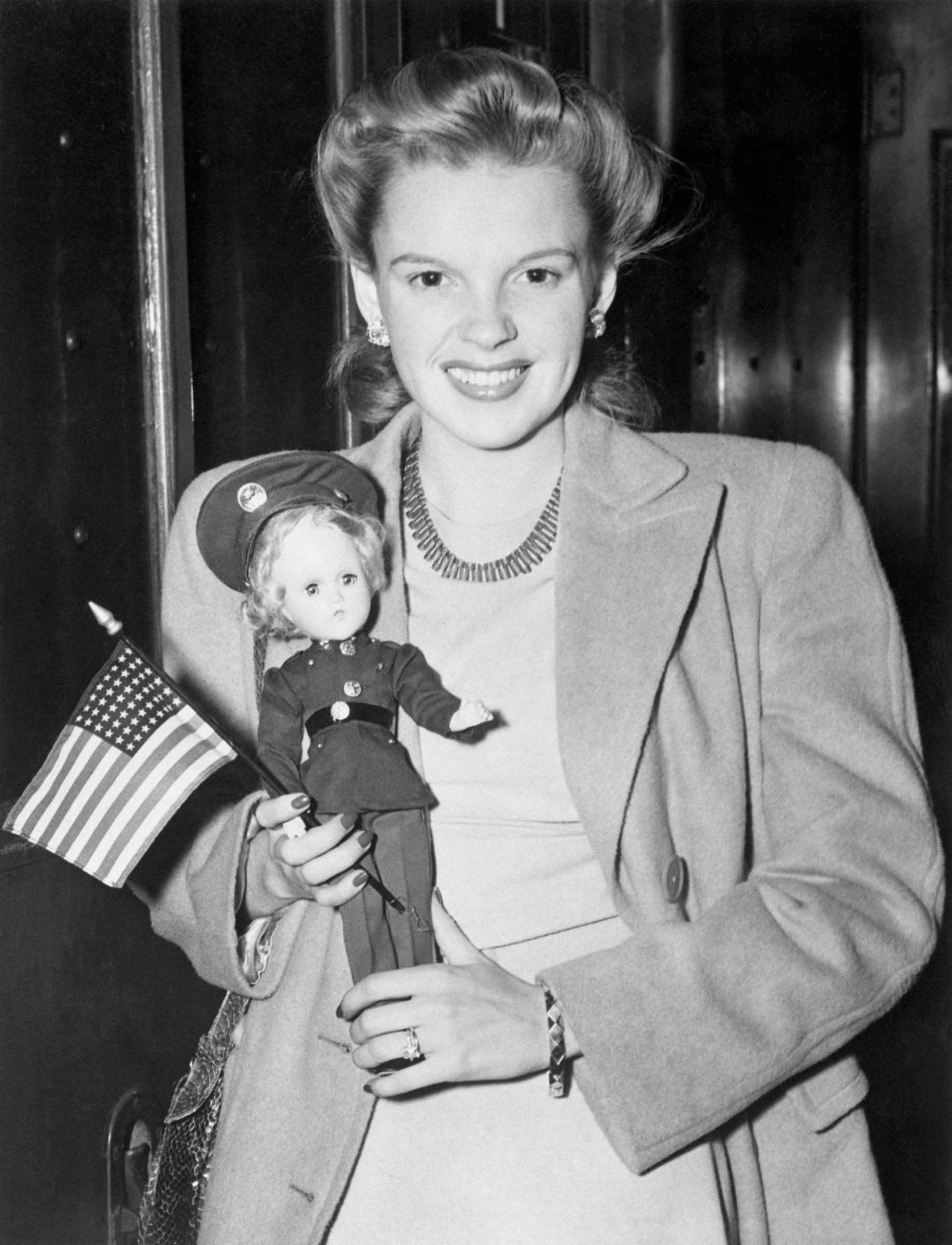 Judy Garland arrive à New York, un drapeau et une poupée à la main, alors qu'elle pose pour les photographes dans une gare | Source : Getty Images