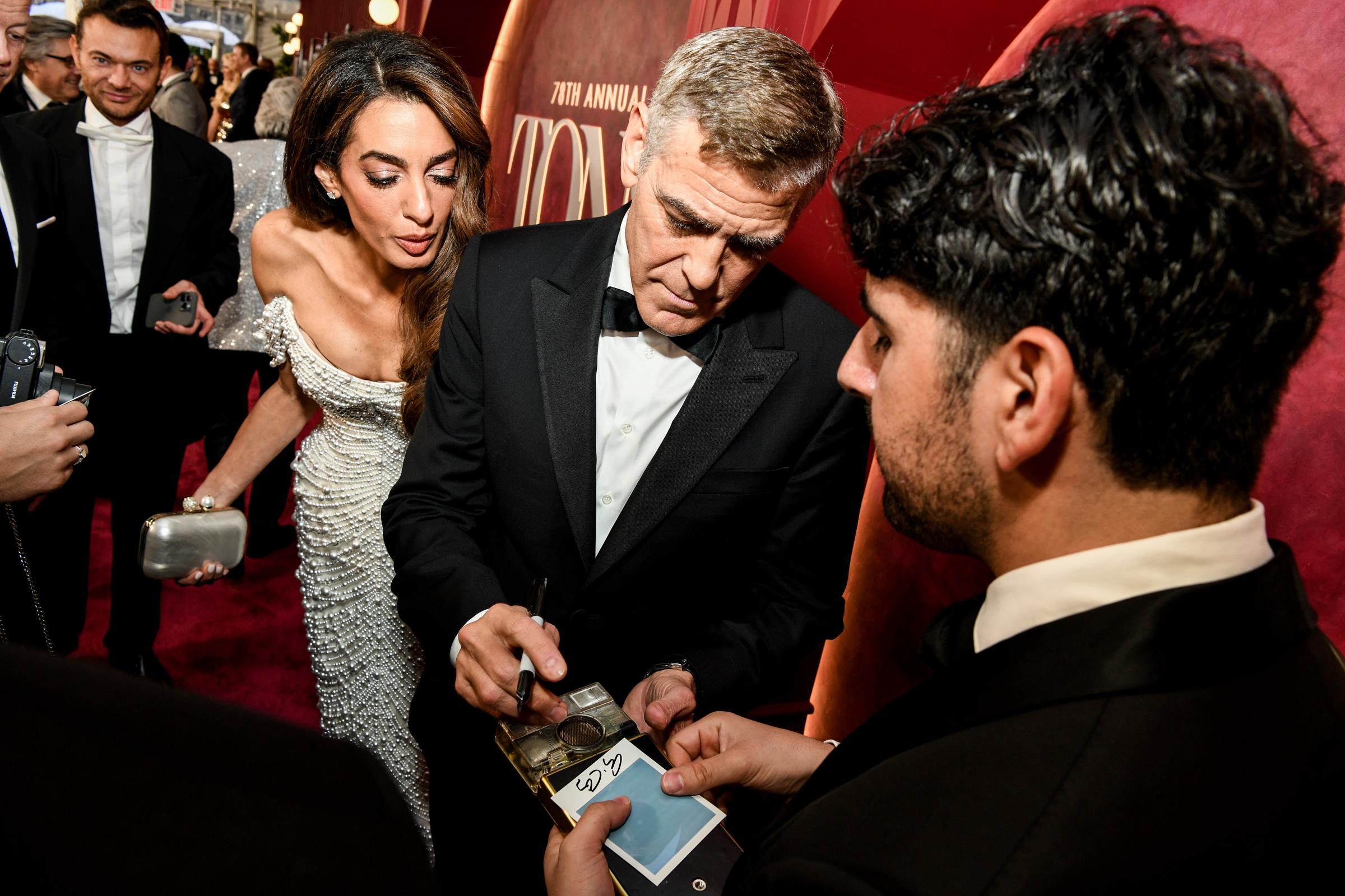 George Clooney signe des souvenirs tandis qu'Amal Clooney se penche pour observer, l'expression concentrée. Le couple fait une pause au milieu d'un tourbillon de fans et de caméras clignotantes sur le tapis rouge.