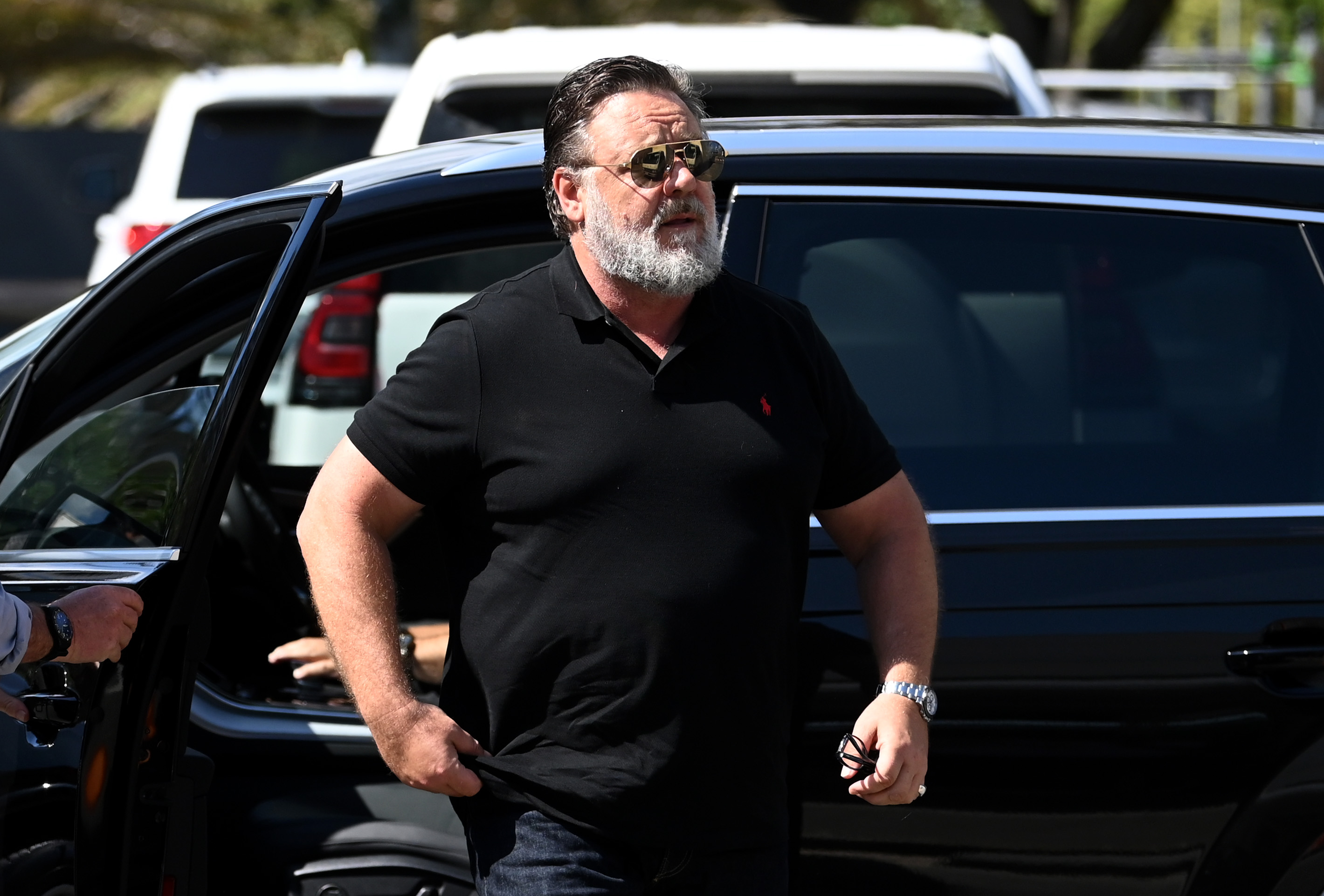 Russell Crowe arrive à une conférence de presse le 28 janvier 2023 à Burleigh Heads, en Australie, vêtu d'un polo noir ajusté et d'un jean. Avec sa barbe blanche, ses lunettes de soleil aviateur et sa carrure imposante, l'acteur semble confiant et décontracté avant la transformation physique qui fera bientôt la une des journaux.