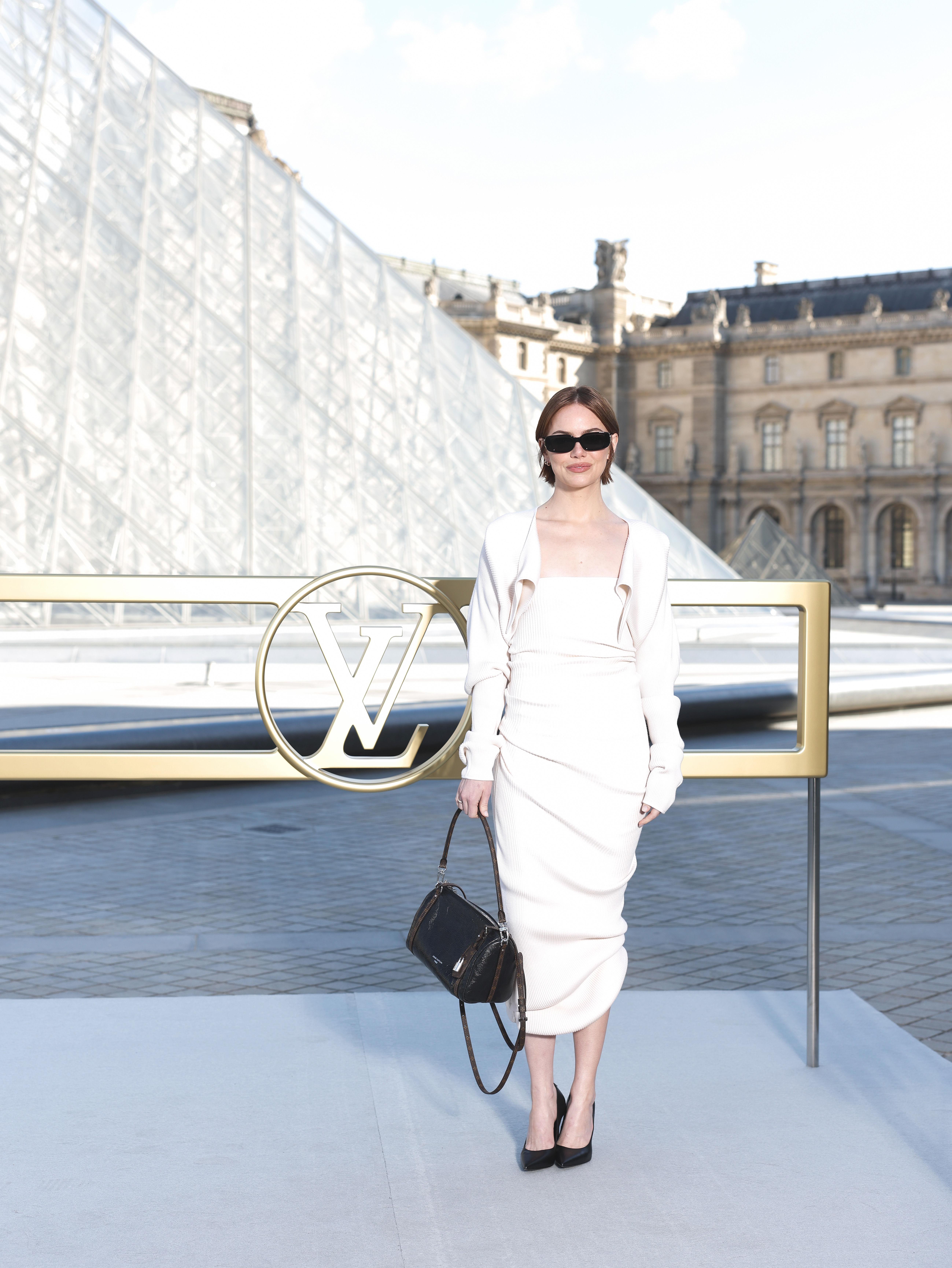 Emma Stone assiste au défilé Louis Vuitton Femme Printemps/Été 2026 lors de la Fashion Week de Paris, en France. Elle porte une robe mi-longue blanc crème, fortement côtelée, qui met en valeur la texture et la silhouette. Elle l'associe à des accessoires noirs élégants pour ancrer le look monochrome.