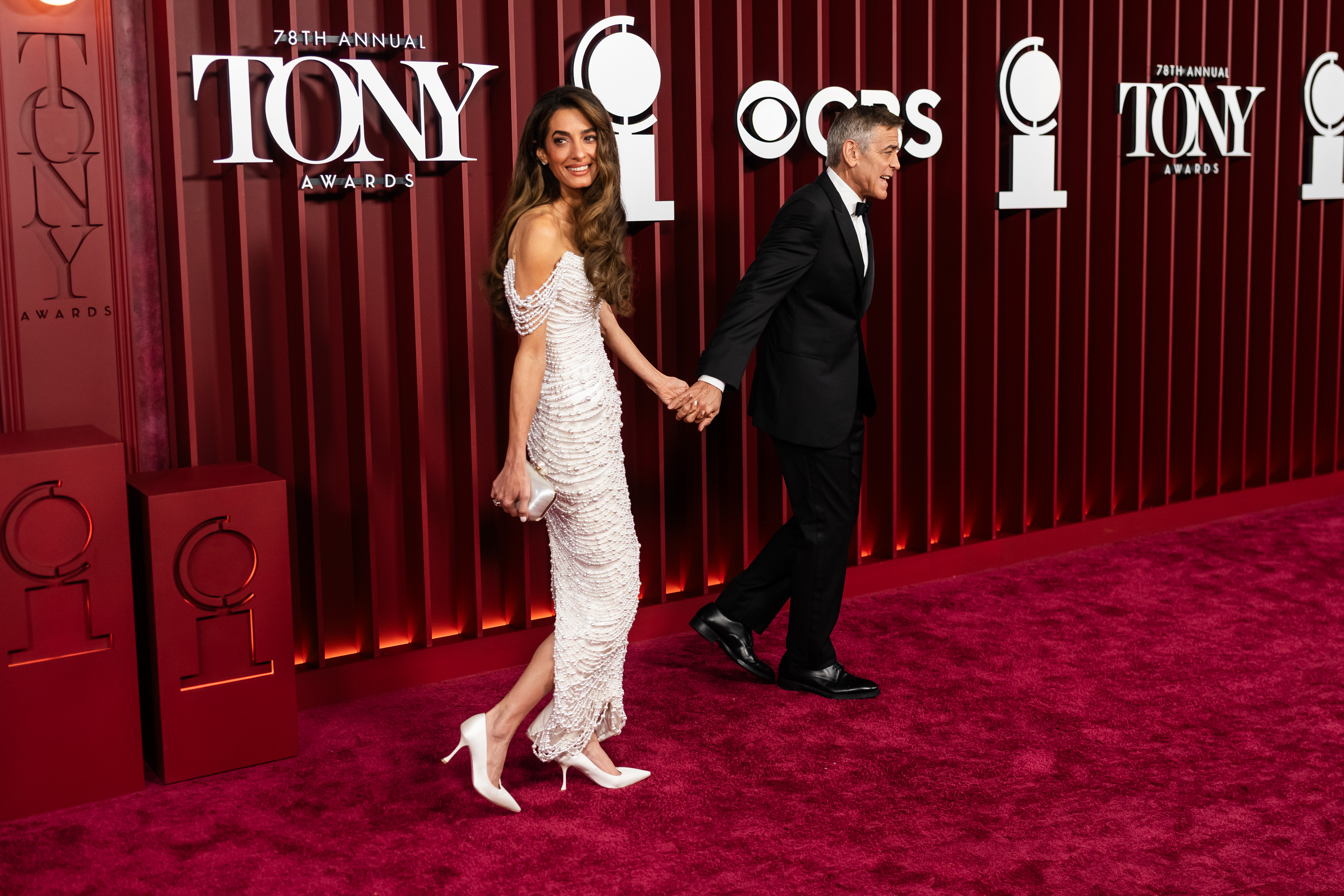 George Clooney conduit Amal Clooney sur le tapis rouge des Tony Awards, le couple échangeant des expressions sous les flashs des appareils photo. La toile de fond caractéristique de l'événement encadre leur arrivée pleine d'aplomb.
