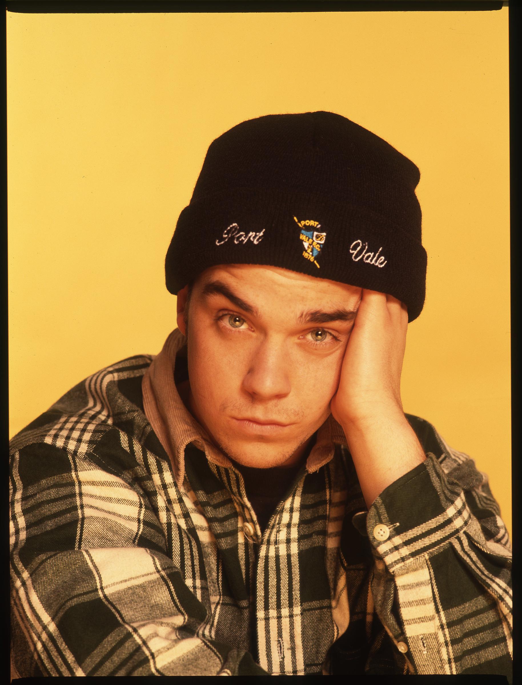 Robbie Williams photographié en mars 1993, la tête appuyée sur sa main, coiffé d'un bonnet Port Vale et vêtu d'une chemise en flanelle verte. L'image capture un rare moment de calme de la part de la star montante, dont le mélange d'attitude et de vulnérabilité séduisait déjà les fans.