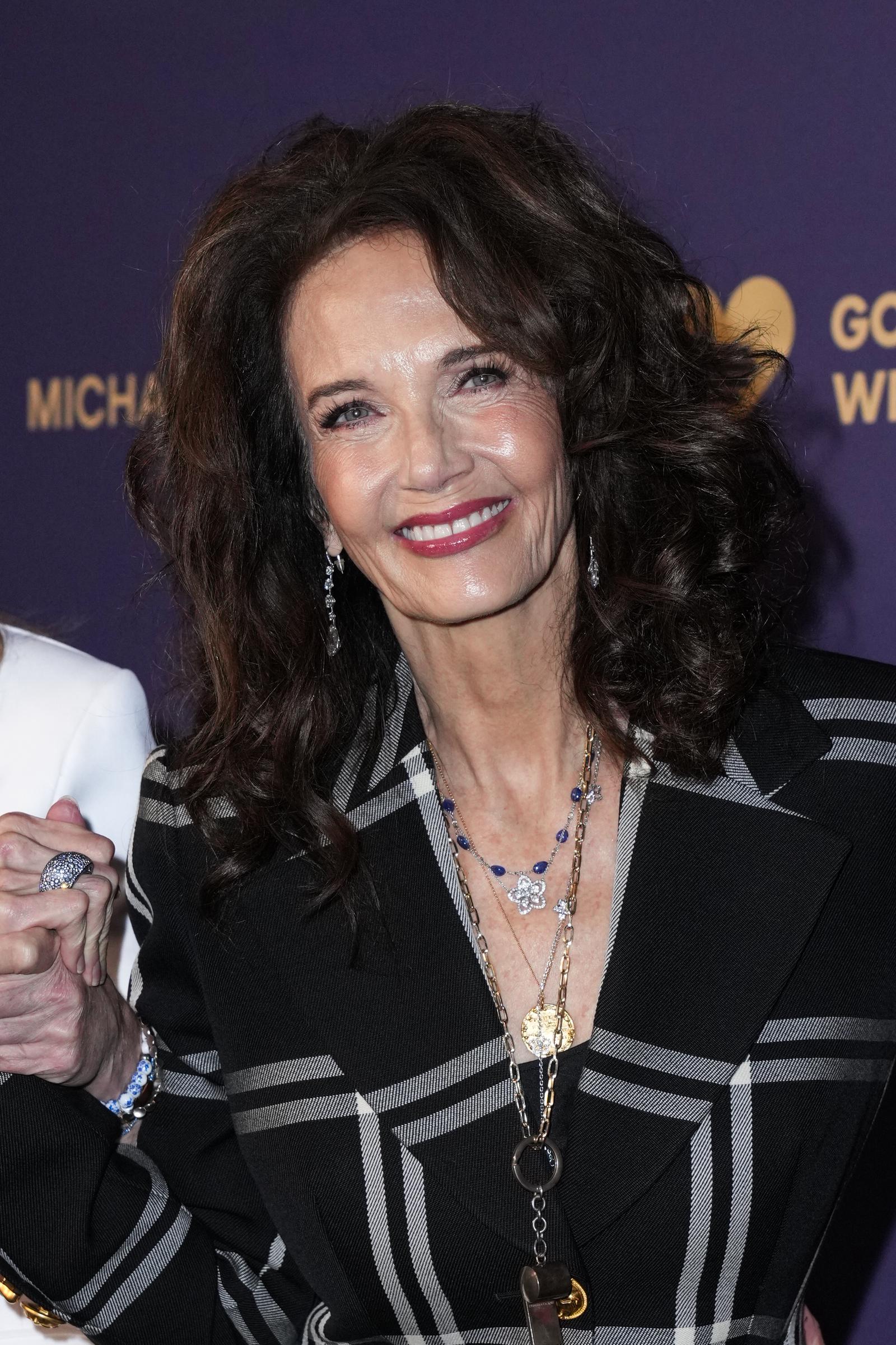 Lynda Carter arbore un sourire radieux sur le tapis rouge de l'événement God's Love We Deliver, vêtue d'un blazer à carreaux noirs et parée de colliers superposés en or et pierres précieuses. Ses boucles volumineuses encadrent son visage, qu'elle accessoirise avec des boucles d'oreilles pendantes et un rouge à lèvres rose vif, dégageant chaleur et charisme.