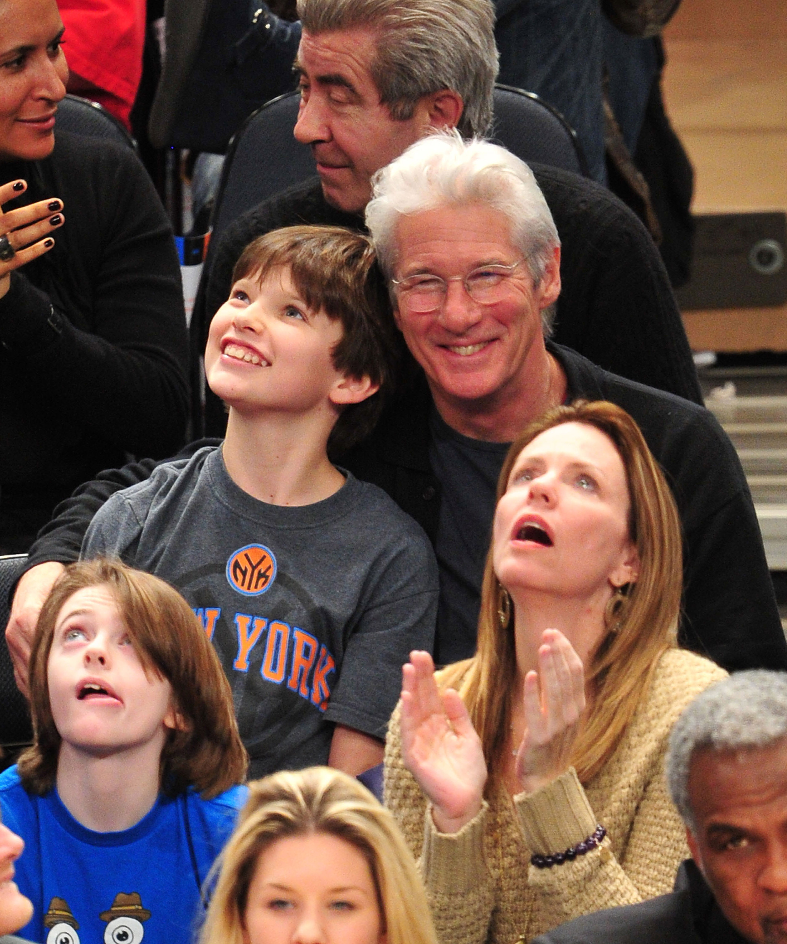 Rayonnant de joie, Richard Gere se penche vers Homer Gere lors d'un match des Knicks au Madison Square Garden de New York, partageant un moment de complicité au bord du terrain, entourés d'autres fans (2012)
