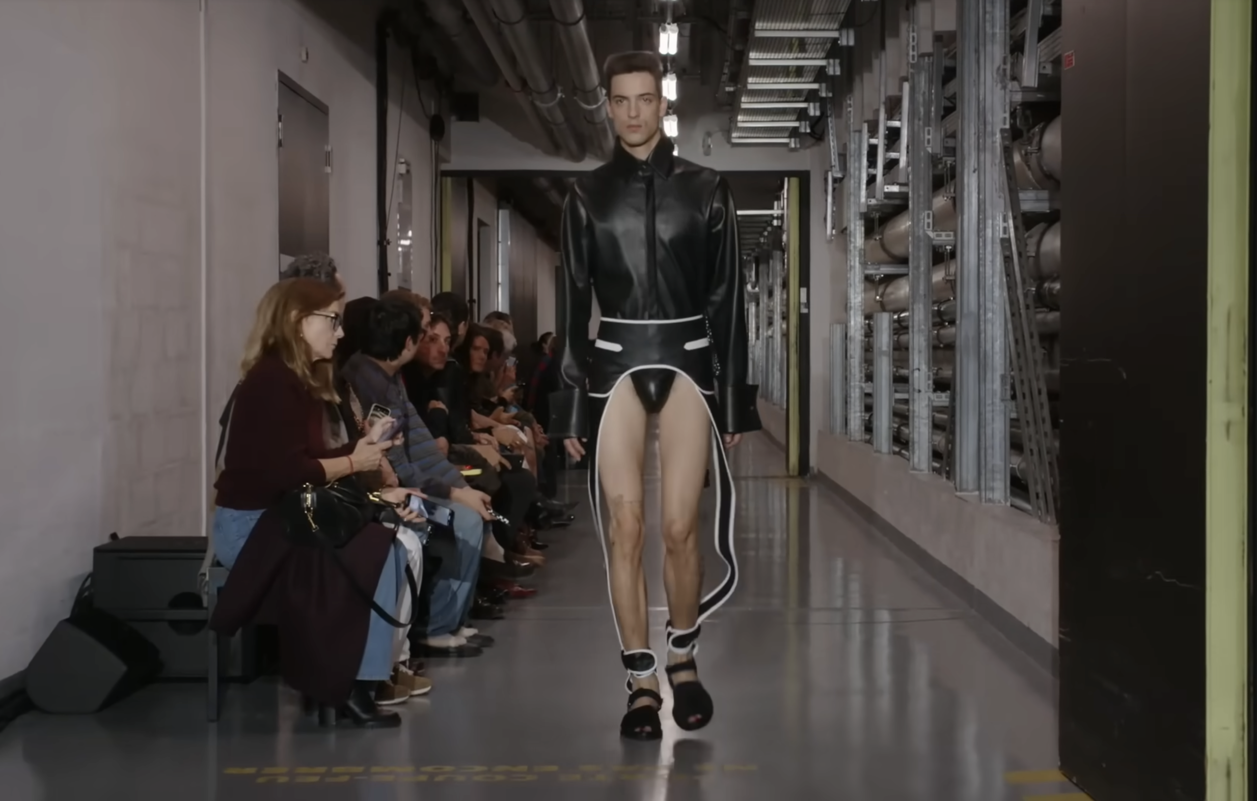 Un mannequin défile dans une tenue noire saisissante qui dévoile l'aine. Le modèle ressemble à un manteau, mais présente des détails latéraux qui descendent le long des jambes, ainsi qu'une section ajustée au centre. Extrait d'une publication datée du 5 octobre 2025. Source : Youtube/@jeanpaulgaultier