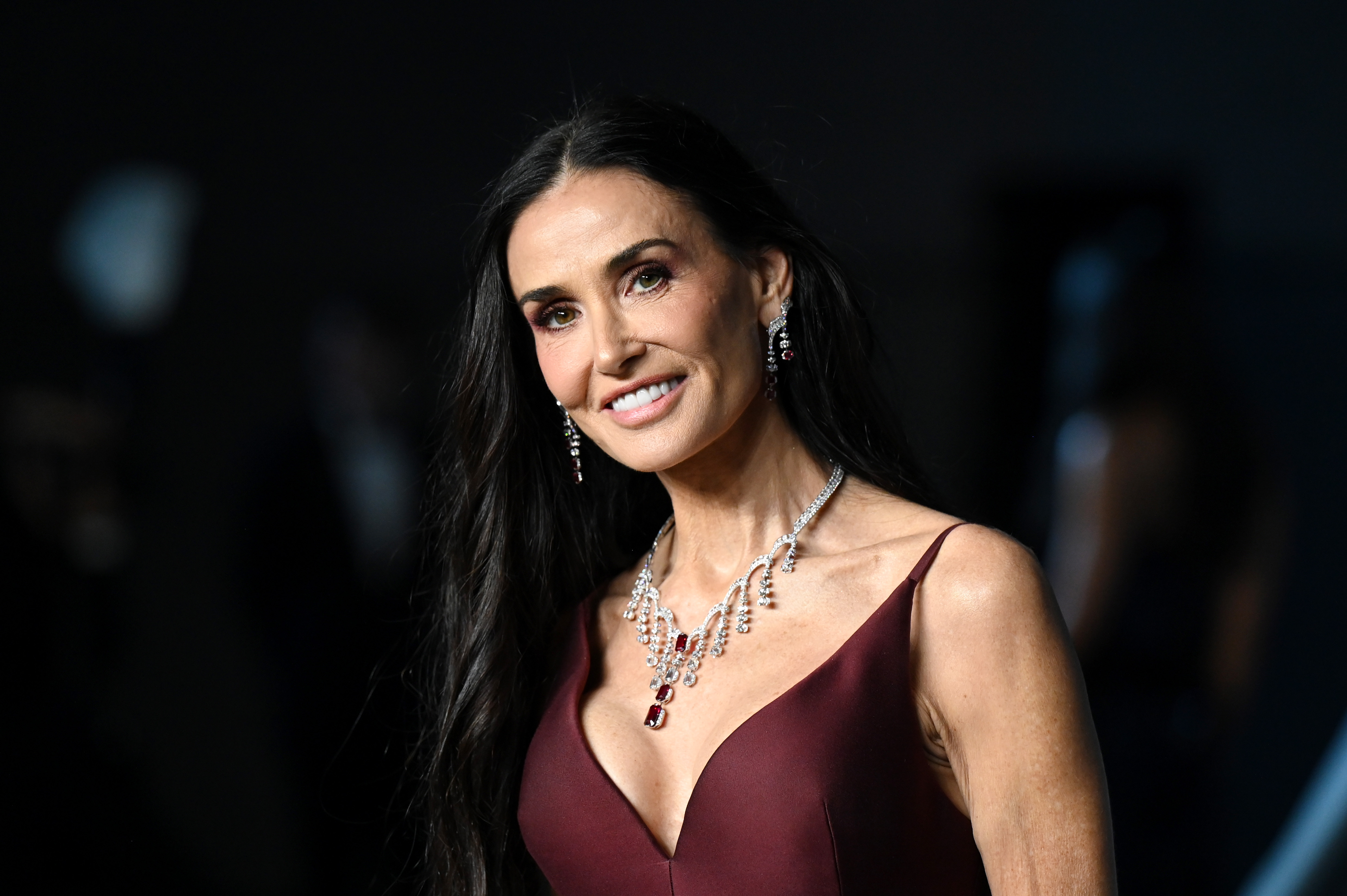 Plan serré sur Demi Moore qui affiche un teint impeccable et un sourire confiant, son collier de rubis étincelant mettant parfaitement en valeur le décolleté plongeant de sa robe bordeaux élégante.