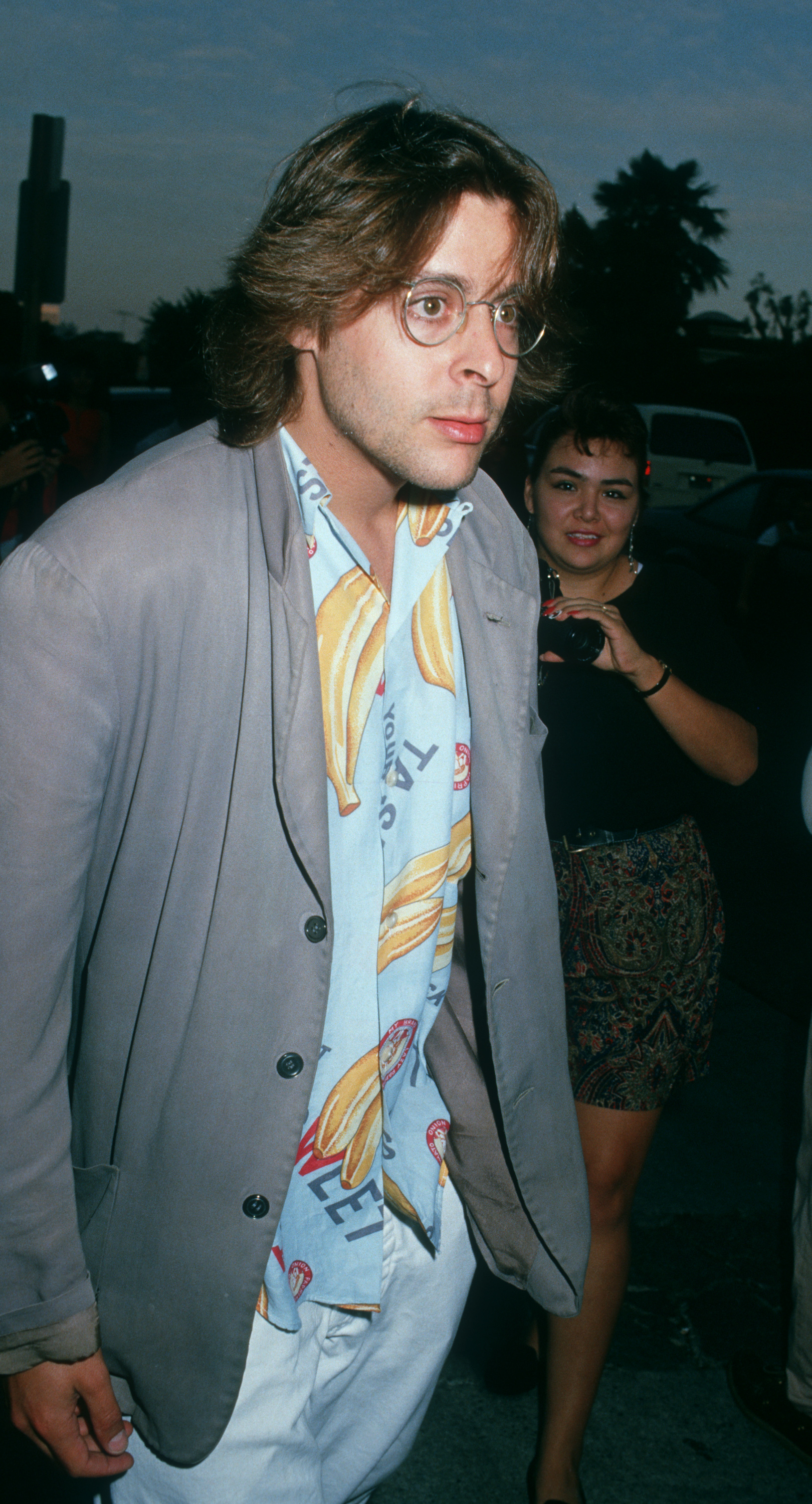Arborant un look décontracté typique de la fin des années 80, avec une chemise à imprimé banane, des lunettes à monture métallique et un blazer gris, l'acteur arrive au cinéma UA Coronet Theater de Westwood pour la première de « Young Guns ». | Source: Getty Images