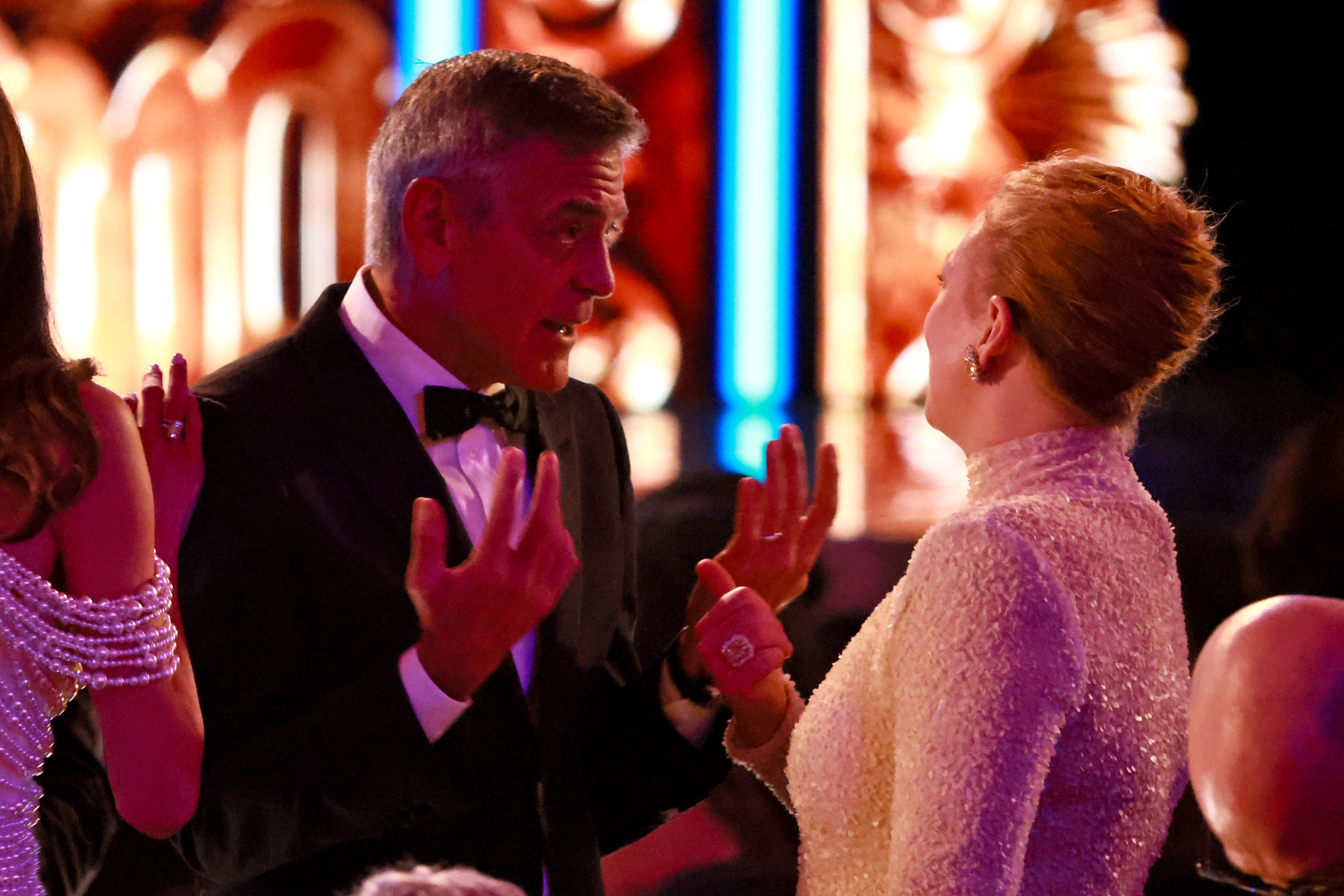 Avec des gestes expressifs, George Clooney discute avec Sarah Snook au milieu du spectacle, tandis qu'Amal Clooney se tient à ses côtés. Le moment animé se déroule dans le théâtre chaleureusement éclairé.