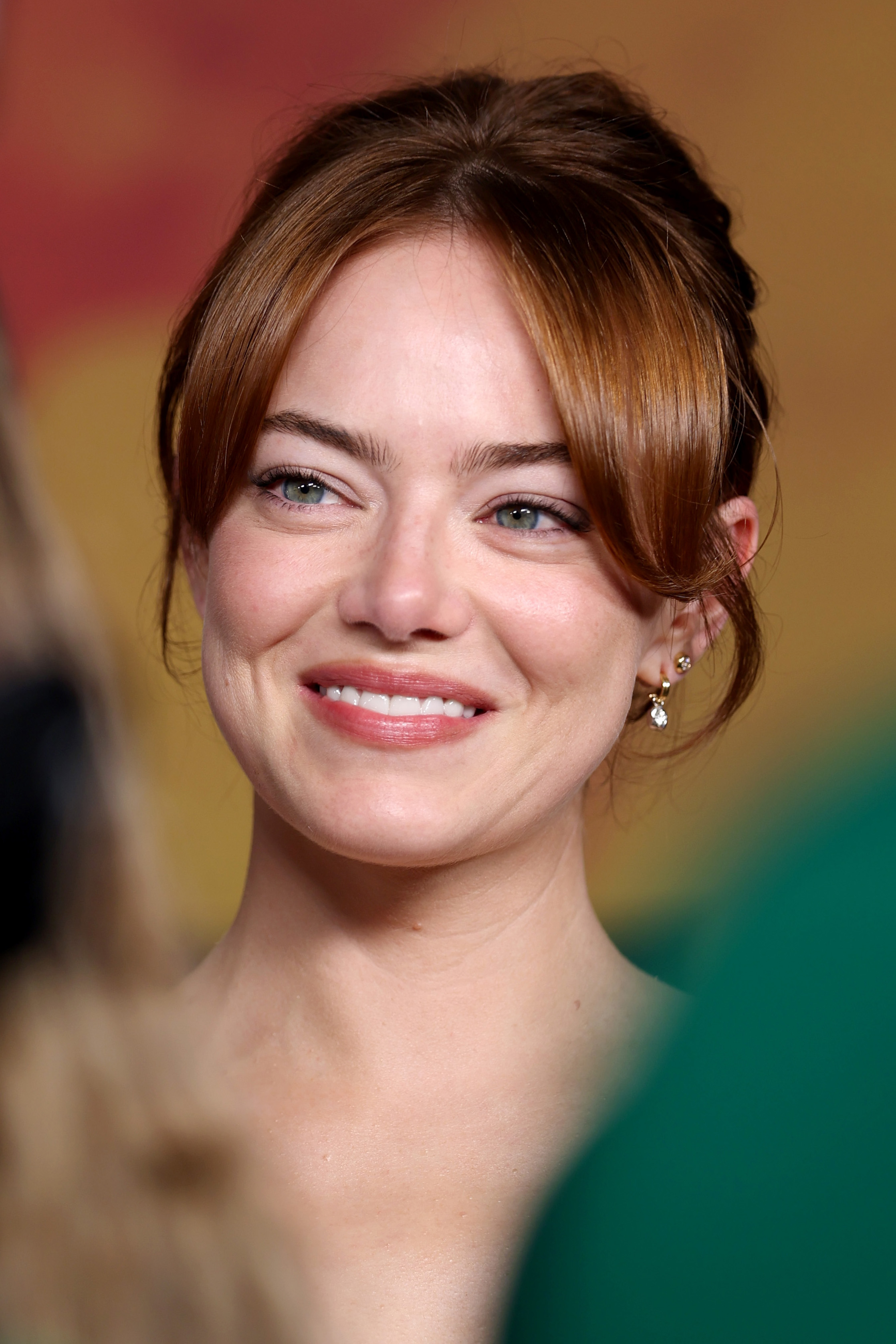 Emma Stone rayonne d'un sourire léger et lumineux, inclinant légèrement la tête tandis que de douces mèches encadrent son visage — une image d'élégance et de confiance tranquille sur le tapis rouge.